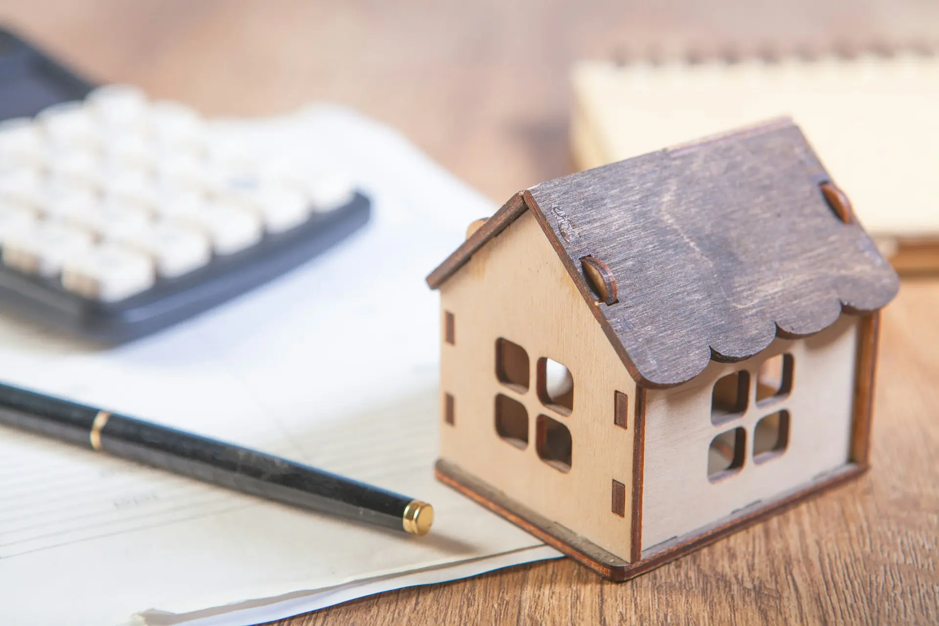 Una casita de madera sobre unos papeles, con una pluma y una calculadora